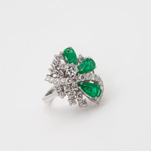 An 18K white gold ring showcasing three pear-shaped emeralds encircled by clusters of small, round diamonds. The design creates a floral or abstract motif, set against a plain white background. Joieria Vintage Barcelona