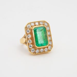 Anillo de oro amarillo 18K con una gran esmeralda rectangular en el centro, rodeada por un borde de diamantes blancos redondos, todo sobre un fondo blanco liso. Joieria Vintage Barcelona