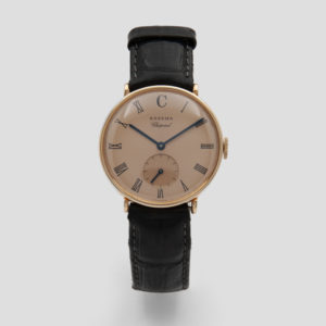 Un reloj de pulsera atemporal con correa de piel negra y caja dorada. La esfera beige muestra números romanos, una subesfera en la posición de las 6 en punto y elegantes manecillas negras. El nombre de la marca "Chopard Eszeha" se muestra elegantemente en la parte superior. Joieria Vintage Barcelona