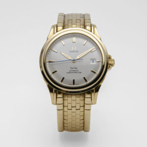 Un rellotge Omega De Ville Co-axial d'or amb esfera de plata, que mostra una finestra de data a les 3 en punt. La polsera metàl·lica millora el seu disseny elegant i clàssic sobre un fons blanc llis. Joieria Vintage Barcelona