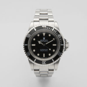 El Rolex Submariner es un reloj de acero inoxidable con esfera y bisel negros. Cuenta con marcadores de hora y manecillas de color blanco, junto con el nombre de la marca y el texto "Submariner" en la esfera. El bisel está marcado con números a intervalos para una medición precisa del tiempo. Joieria Vintage Barcelona