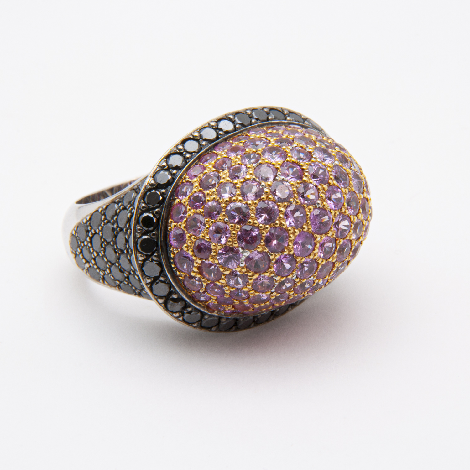 El anillo de oro blanco de 18 quilates con zafiros rosas y diamantes negros presenta un diseño lujoso con una gran parte superior ovalada engastada con numerosos zafiros rosados pequeños. La banda oscura y detallada crea un elegante contraste con las piedras vibrantes, bellamente resaltadas contra el fondo blanco liso. Joieria Vintage Barcelona