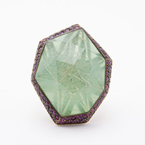 The Vasari ring, crafted in 18K rose gold, features a large rutilated quartz gemstone with a complex internal pattern set within a hexagonal frame. The setting is adorned with small rubies and turquoise accents, creating an intricate and striking design. Joieria Vintage Barcelona