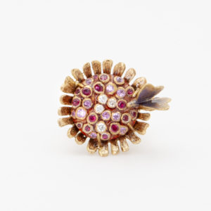 El "Anillo Vasari de oro rosa de 18 quilates" presenta un diseño de inspiración vintage que se asemeja a una flor, con una disposición circular de rubíes, zafiros rosados y diamantes en el centro. Un delicado detalle similar a una pluma se extiende desde un costado, que recuerda a un pétalo, lo que le suma un atractivo ornamental. Joieria Vintage Barcelona