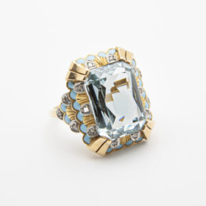 Aquest és un anell luxós Art Deco d'or groc de 18 quirats que presenta una pedra preciosa aiguamarina sintètica rectangular de grans dimensions encastada enmig d'un disseny intricat, ressaltat amb petits diamants circulars i detalls d'esmalt blau clar. Joieria Vintage Barcelona