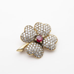An 18K yellow gold brooch with ruby ​​and diamonds, in the shape of a four-leaf clover, features a design encrusted with small diamonds and a central ruby ​​on a white background. Vintage Jewelry Barcelona