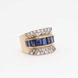 Us presentem l'Anell anys 40´d´or groc 18K amb safirs i diamants: un impressionant anell d'or adornat amb una banda central de safirs blaus rectangulars, flanquejat amb elegància per grups de petits diamants blancs a cada costat, tot sobre un fons blanc senzill. Joieria Vintage Barcelona