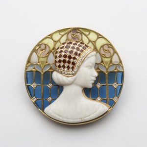 El "Broche-medalla novecentista de Masriera" es un broche Art Déco de oro amarillo de 18 quilates que presenta un relieve de perfil lateral del rostro de una mujer, sobre un fondo circular de esmalte plique-à-jour con motivos azules y verdes. La mujer está adornada con un tocado decorativo adornado con joyas y rubíes. Joieria Vintage Barcelona