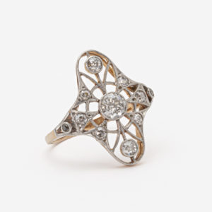 Un detallat anell d'estil vintage anomenat "Anell Belle Epoque", elaborat amb or de 18 quilates, platí i diamants, presenta un disseny semblant a un encaix amb diversos diamants petits i un diamant central més gran en una banda de plata, elegantment mostrat sobre un fons blanc llis. Joieria Vintage Barcelona