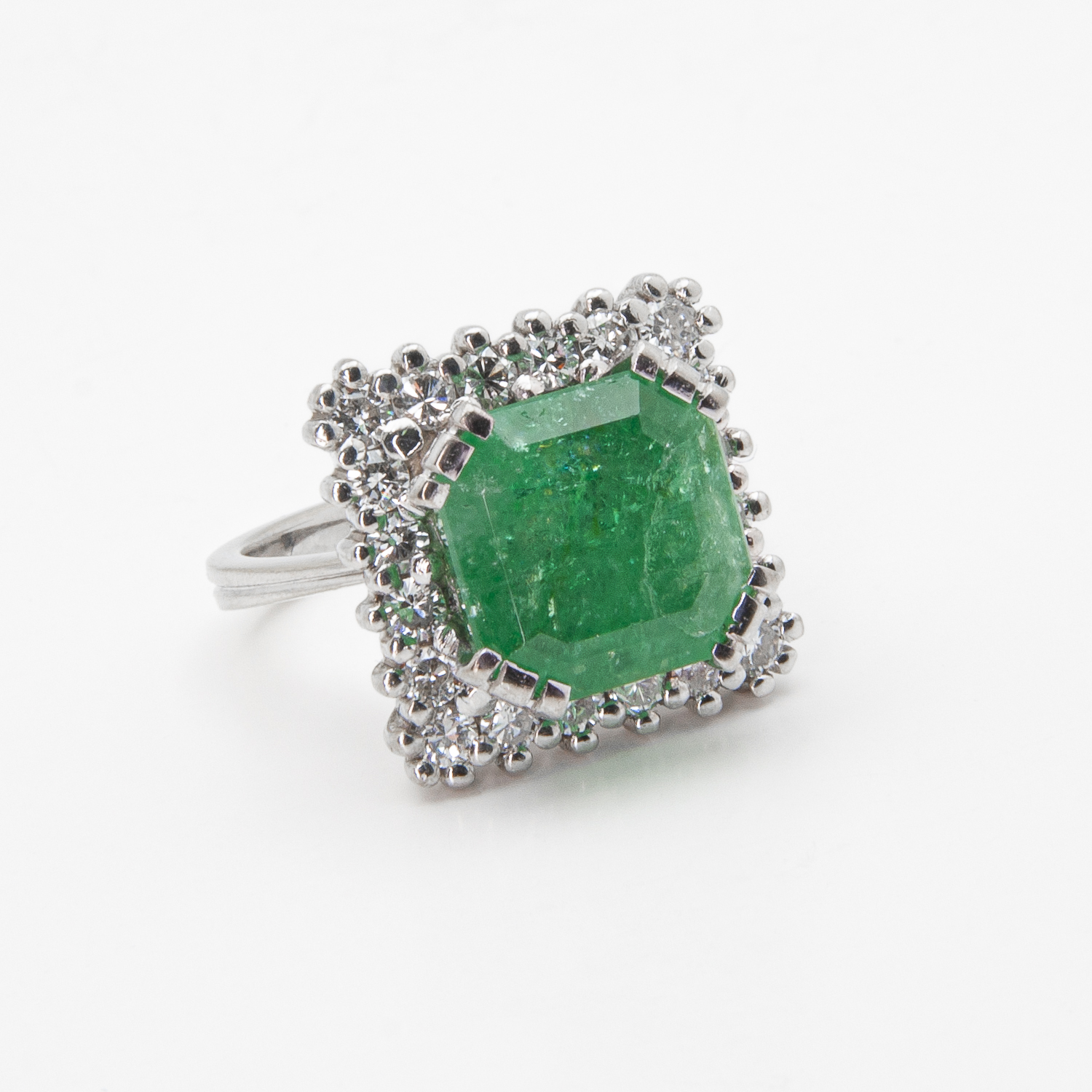 El Anillo de oro blanco de 18 quilates con esmeralda y diamantes exhibe una prominente esmeralda verde de corte cuadrado engastada en plata, rodeada por un halo de pequeños diamantes redondos, todos presentados sobre un sencillo fondo blanco. Joieria Vintage Barcelona