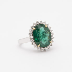 Un anell de plata que mostra una gran pedra preciosa ovalada de color verd està envoltat per un halo de petits diamants rodons, tots encaixats sobre un fons blanc llis, es descriu com l'"Anell d'or blanc de 18K amb maragda i diamants. Joieria Vintage Barcelona