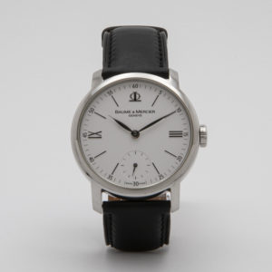 Primer pla del Baume & Mercier Classima acer, un elegant rellotge de polsera analògic, que mostra la seva esfera rodona blanca amb números romans negres i una petita subesfera a la posició de les 6 en punt. El rellotge compta amb una corretja de cuir negra i està revestit de plata, amb la inscripció "Baume & Mercier" en un lloc destacat a l'esfera. Joieria Vintage Barcelona