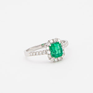 Un anell de plata amb una maragda verda de tall quadrat envoltada de petits diamants, acompanyat d'una banda adornada amb diamants a banda i banda de la peça central, es mostra elegantment sobre un fons blanc llis. Aquest producte s'anomena "Anell d'or blanc 18K amb maragda i diamants". Joieria Vintage Barcelona