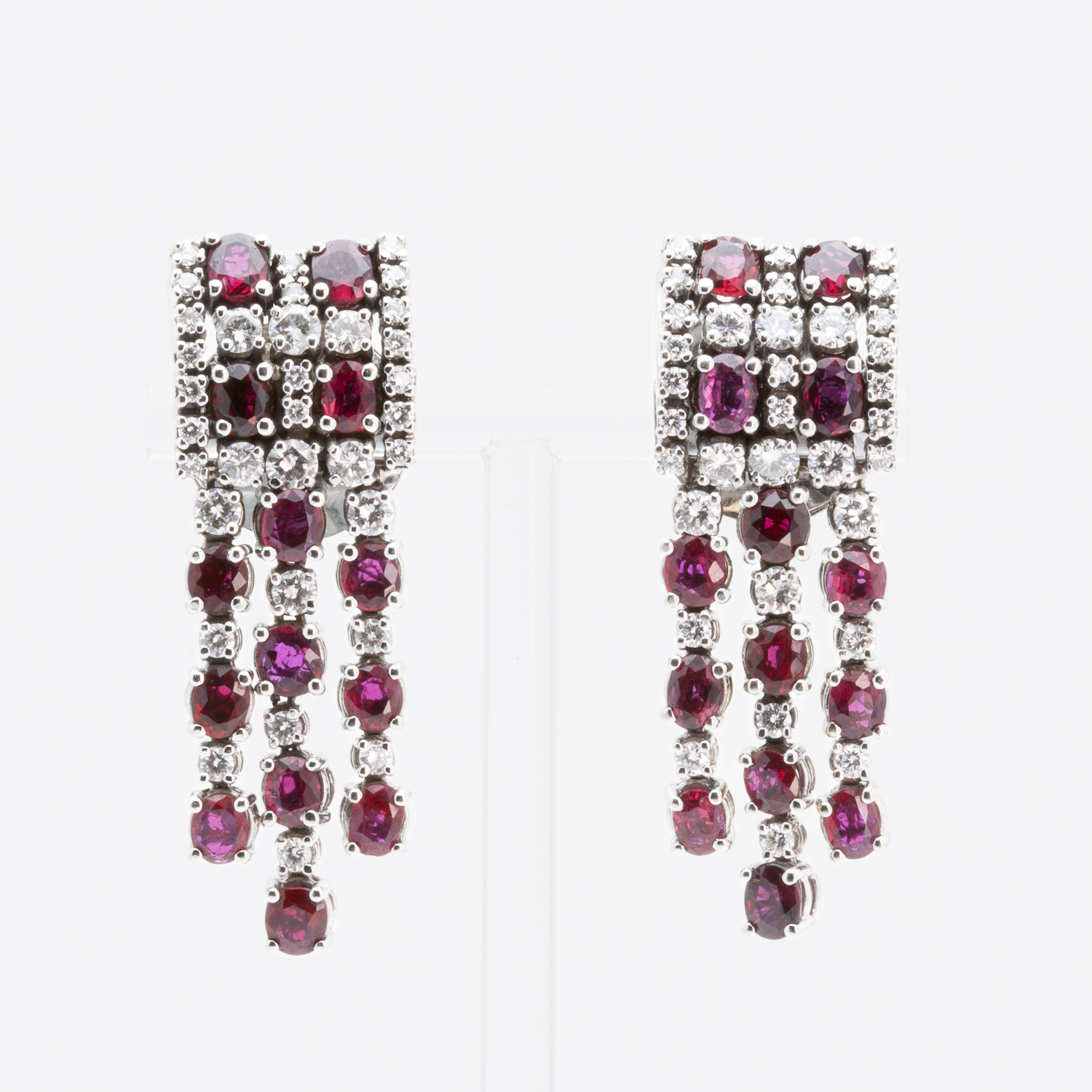 Un par de sofisticados aretes de oro blanco de 18 quilates con diamantes y rubíes, que presentan tres tiras colgantes de piedras preciosas rojas, sobre un fondo blanco liso. Joieria Vintage Barcelona