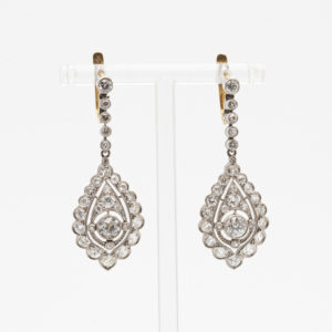 Los pendientes Belle Epoque de oro, platino y diamantes presentan un exquisito diseño en forma de pera en el centro, adornado con numerosos diamantes redondos brillantes engastados en un patrón simétrico. Los pendientes cuelgan con gracia de una hilera más pequeña de gemas que se conectan a un gancho. Joieria Vintage Barcelona