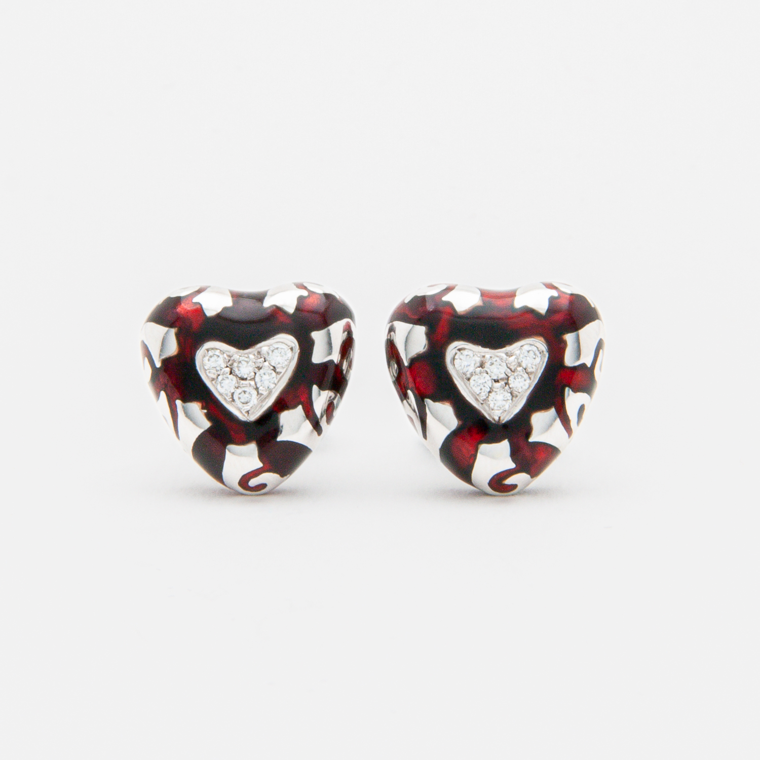 A pair of "Fidia" earrings crafted in 18-karat white gold, featuring heart-shaped designs adorned with spirals of black and red enamel. Each earring showcases a central heart embellished with inlaid diamonds, set against a smooth white background. Vintage Jewelry Barcelona