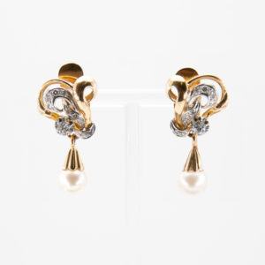 A pair of gold and platinum earrings from the 1940s featuring intricate spiral designs, adorned with diamonds and a dangling pearl at each end, displayed on a transparent stand against a plain background. Vintage Jewelry Barcelona