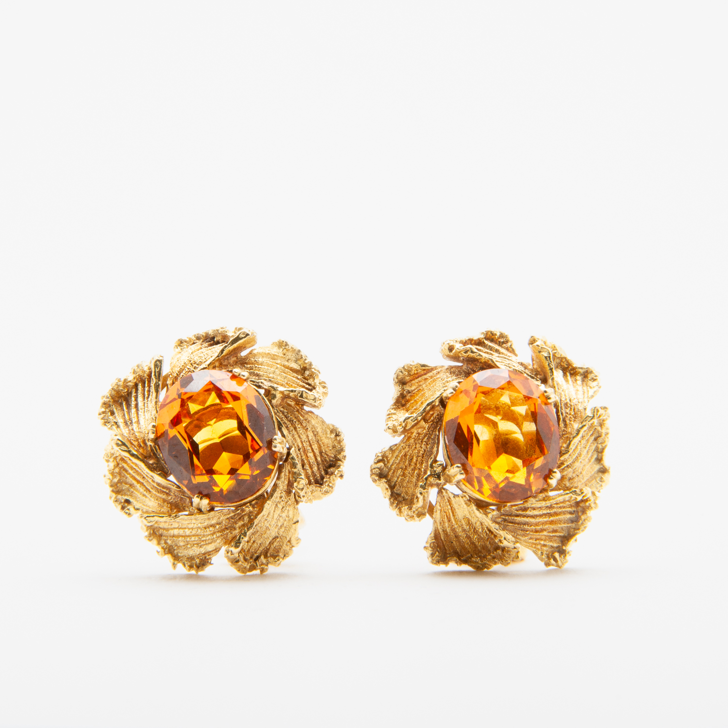 Les Arracades d'or i safir taronja són arracades d'or de disseny intricat amb pedres precioses d'ambre centrals, que s'assemblen a pètals florals al voltant de les pedres sobre un fons blanc llis. Joieria Vintage Barcelona