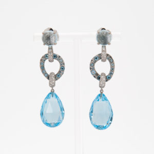 Los pendientes Vasari en oro blanco de 18 quilates presentan elegantes aros de plata colgantes adornados con piedras preciosas de topacio azul y diamantes, cada uno de los cuales sostiene una gran gema facetada en forma de lágrima azul, todo ello exhibido en un soporte transparente sobre un fondo sencillo. Joieria Vintage Barcelona