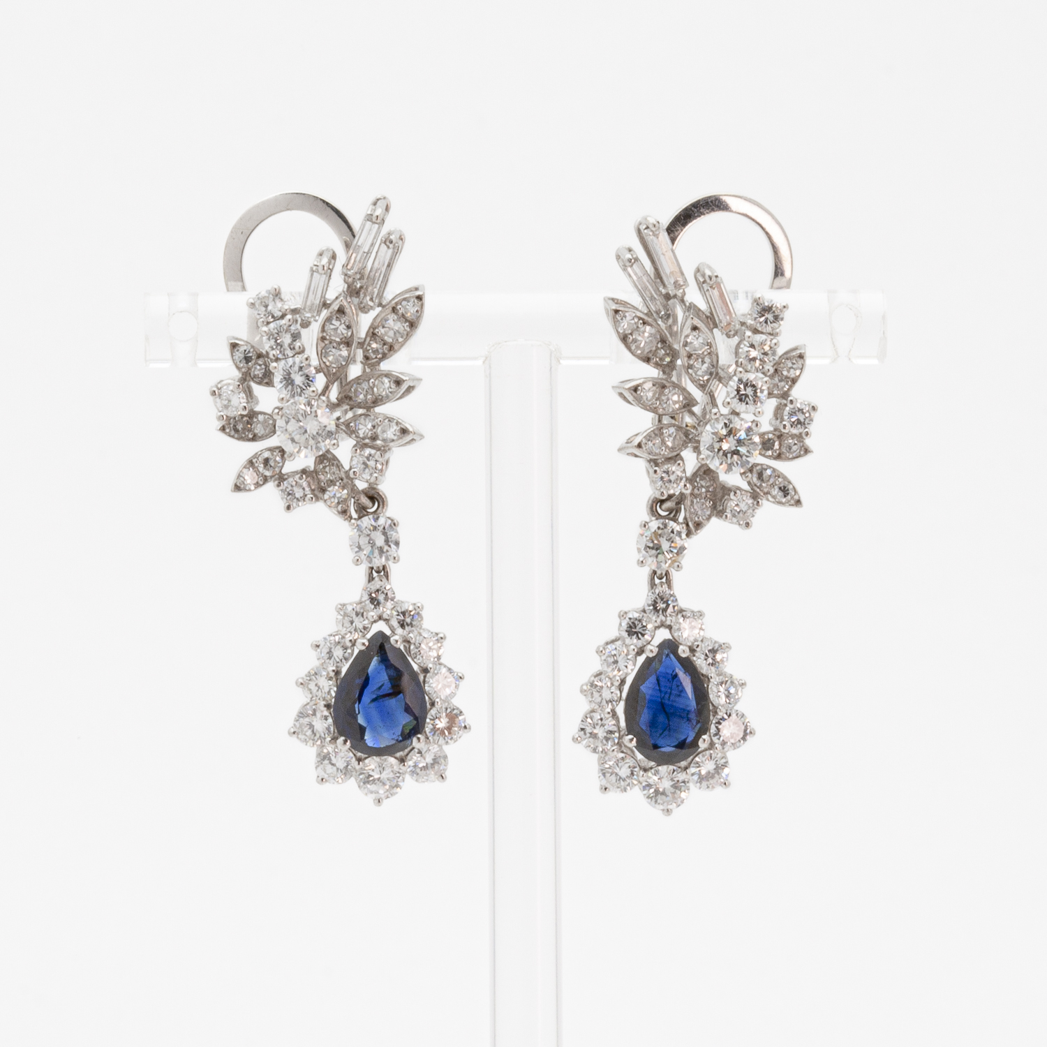 The "three-in-one" white gold earrings feature a floral design with clusters of clear gemstones, centered around a prominent teardrop-shaped blue gemstone surrounded by smaller stones. Elegantly presented on a white setting, they incorporate diamonds, rubies, and sapphires. Vintage Jewelry Barcelona