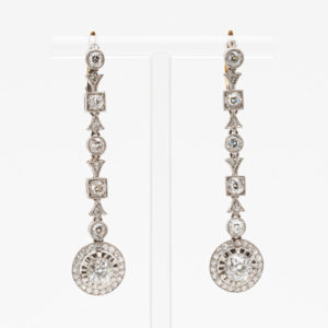Par de elegantes pendientes colgantes con engastes de diamantes circulares y cuadrados, cada uno con un gran diamante redondo en la parte inferior. Los pendientes se presentan en un soporte transparente sobre un fondo blanco. Joieria Vintage Barcelona