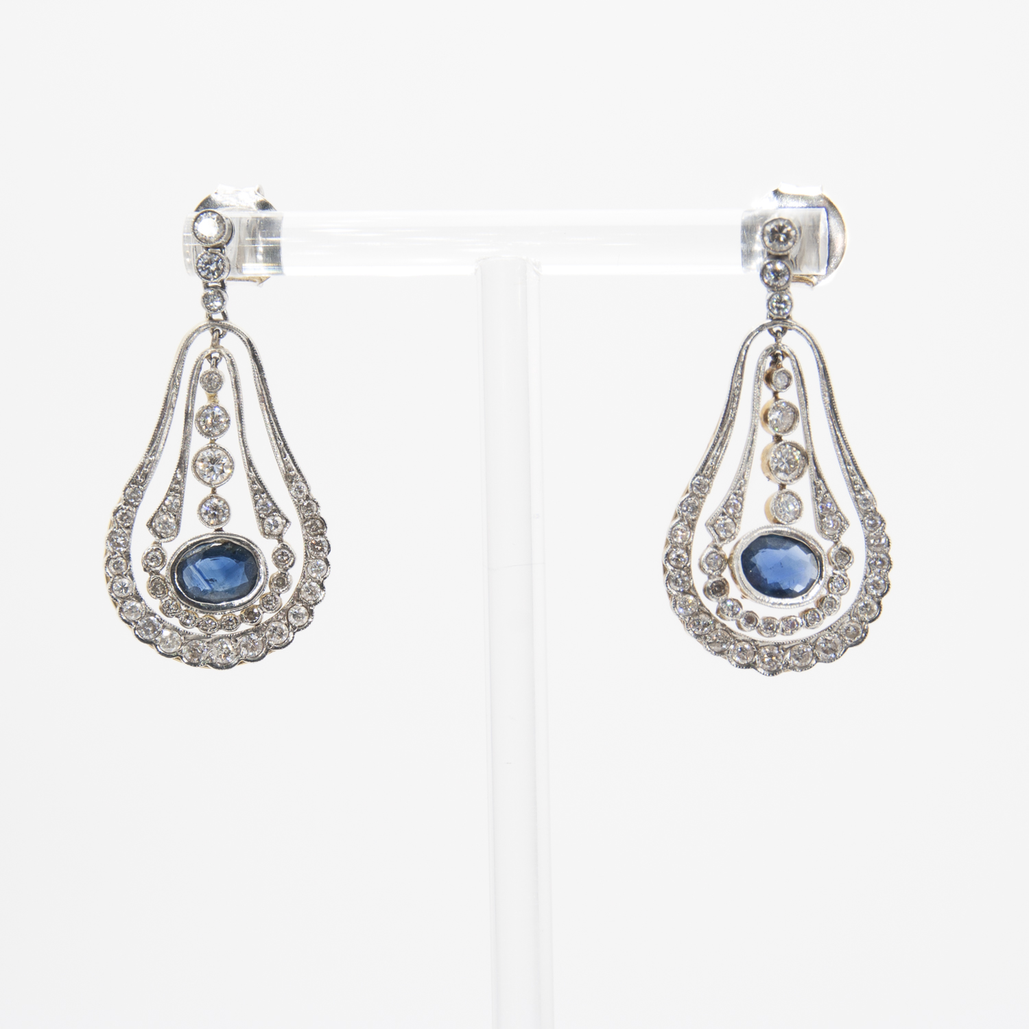 Estos elegantes pendientes en forma de lágrima presentan intrincadas bandas de plata con brillantes piedras preciosas blancas que rodean una piedra azul ovalada central, que se exhibe en un soporte transparente. Estos son los impresionantes "Pendientes estilo Belle Epoque con zafiros y diamantes". Joieria Vintage Barcelona