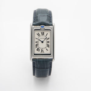 El Cartier Basculant és un rellotge de luxe amb caixa rectangular platejada, esfera blanca amb números romans negres i agulles blaves, i corretja de pell texturitzada gris fosc. També inclou un capbussó blau a la corona. Joieria Vintage Barcelona