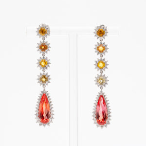Los "Pendientes con zafiros padparadscha, diamantes y zafiros amarillos" presentan elegantes aretes colgantes con grandes piedras preciosas en forma de lágrima, adornados con piedras redondas más pequeñas en un patrón floral y cristales brillantes. Joieria Vintage Barcelona
