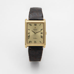 El Rolex Cellini Ref. 4105 presenta una caixa rectangular daurada, una esfera color crema amb números romans negres, dues agulles i la inscripció "Cellini" a l'esfera. Una corretja de pell negra texturitzada complementa el disseny sobre un fons blanc llis.   Joieria Vintage Barcelona