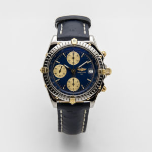 El Breitling Chronomat és un rellotge de polsera de luxe amb esfera blava i detalls daurats, tres subesferes, cronògraf i data. Incorpora una corretja de pell negra amb repunts blancs. Joieria Vintage Barcelona