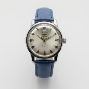 El Longines Conquest Heritage presenta una cara rodona analògica platejada, marcadors d'hora daurats, una finestra de data a les 12 en punt i una corretja de cuir blau tot sobre un fons blanc llis. Joieria Vintage Barcelona