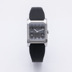 El Baume Mercier Hampton Lady és un rellotge de polsera rectangular amb esfera negra, números platejats, bisell encastat amb diamants i corretja negra, sobre un fons blanc. Joieria Vintage Barcelona
