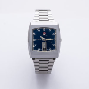 El Rado Diastar-Longines és un rellotge de polsera platejat amb una esfera blava quadrada, marcadors d'hora platejats, corretja de baules de metall, indicador de dia i data, i la marca "Rado" a l'esfera. Joieria Vintage Barcelona