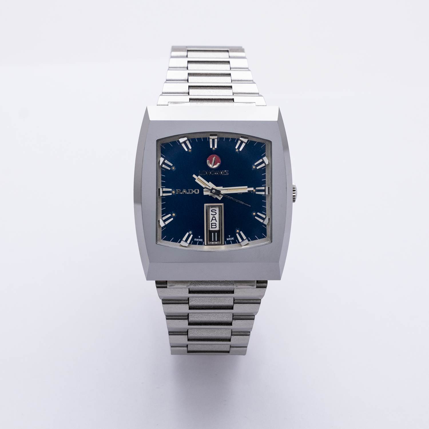 El Rado Diastar-Longines es un reloj de pulsera plateado con una esfera azul cuadrada, marcadores de hora plateados, correa de eslabones de metal, indicador de día y fecha, y la marca "Rado" en la esfera. Joieria Vintage Barcelona