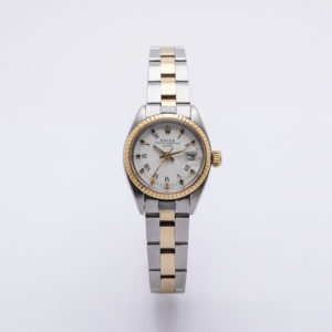 El Rolex Lady Oyster Perpetual Date és un rellotge de polsera de luxe que compta amb una corretja de metall de dos tons plata i or, una esfera blanca rodona amb números romans, bisell daurat, finestra de data i la marca “Rolex” visible a la cara. Joieria Vintage Barcelona