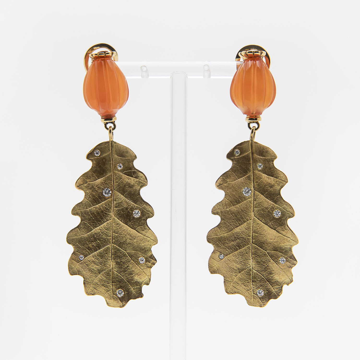 Los Pendientes Vasari de oro 18k, con diamantes y cornalina son pendientes colgantes con remates de cuentas naranjas y grandes colgantes dorados en forma de hoja adornados con piedras preciosas, que se muestran sobre un soporte transparente sobre fondo blanco. Joieria Vintage Barcelona