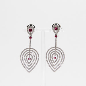 Los Pendientes Vasari son elegantes pendientes colgantes de oro blanco de 18 quilates con aros concéntricos en forma de lágrima adornados con diamantes y acentuados con rubíes, que se exhiben en un soporte transparente sobre fondo blanco. Joieria Vintage Barcelona
