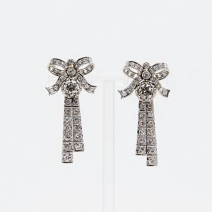 Pendientes Belle Époque de platino y diamantes: elegantes pendientes en forma de lazo, decorados con diamantes redondos y cuadrados, con dos tiras colgantes bajo la gema central. Presentados sobre fondo blanco. Joieria Vintage Barcelona